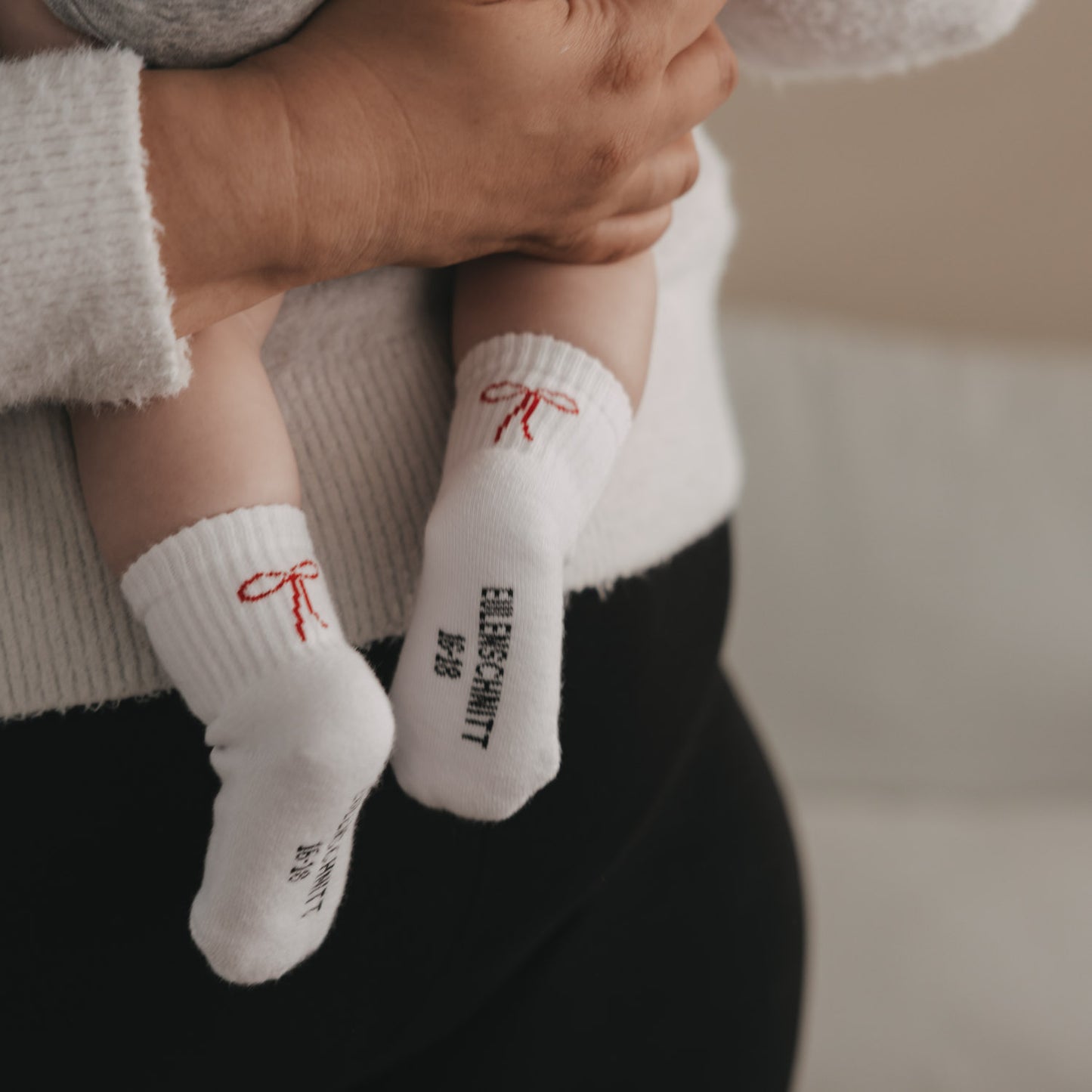 Baby auf dem Arm mit weißen Socken und roter Schleife, Eulenschnitt Schriftzug sichtbar