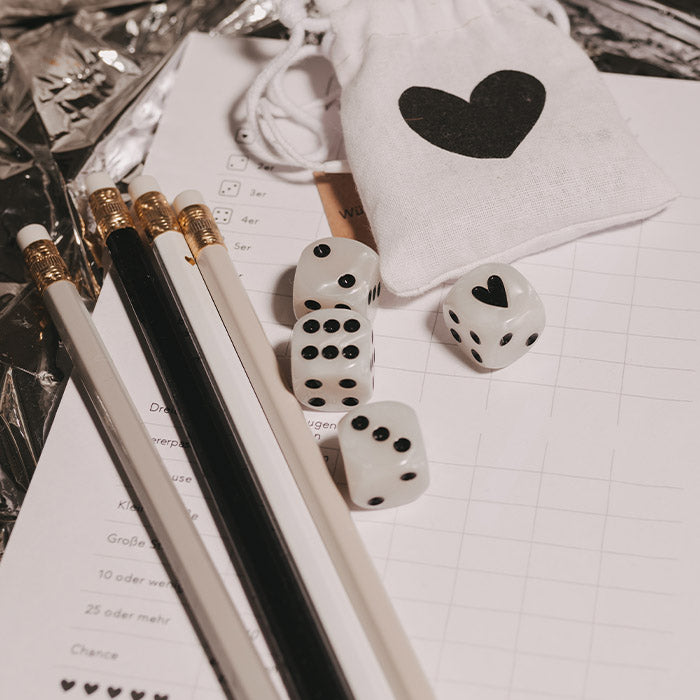 Perlmuttfarbene Würfel mit schwarzen Punkten und Herzmotiv, zusammen mit schwarz-weißen Stiften, Spielblock und einem weißen Stoffbeutel mit schwarzem Herz.