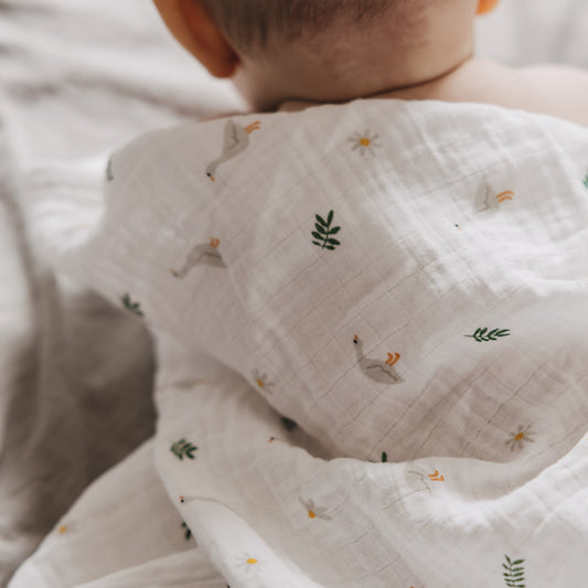 Baby liegt auf einer weichen Unterlage und ist in ein leichtes Musselintuch mit Gänsefamilien-Muster eingehüllt. Die sanften Farben und verspielten Motive schaffen eine liebevolle Atmosphäre.