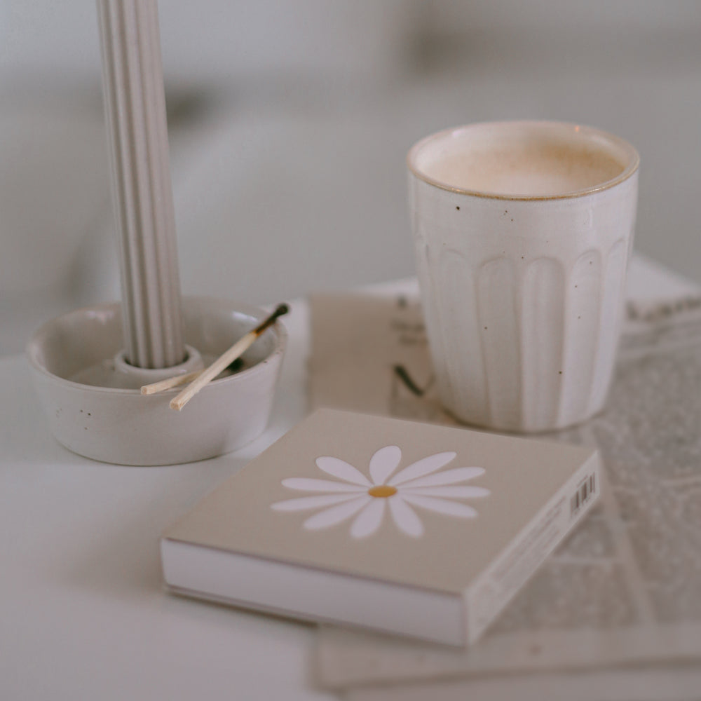 Beige Streichholzschachtel Daisy mit Blumenmotiv neben Kerzenhalter und Tasse – harmonisch inszeniert.