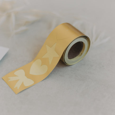 Goldene Aufkleberrolle mit Motiv Stern, Herz, Schleife auf hellem Tisch mit Trockenblumen