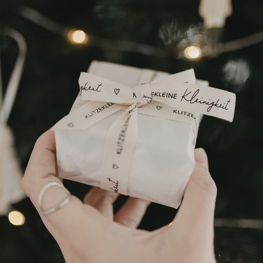 Hand hält Geschenk mit Baumwollband „Klitzekleine Kleinigkeit“ vor weihnachtlichem Hintergrund