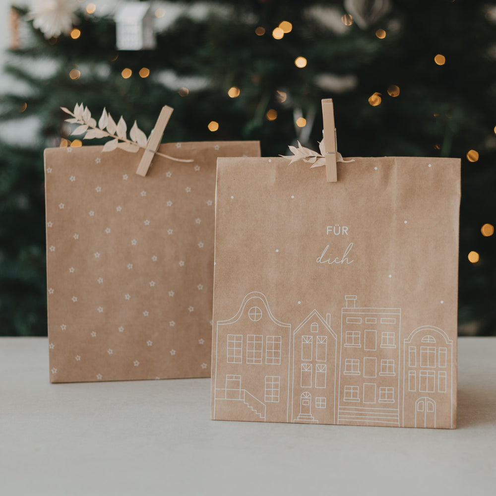 Geschenktüte aus Kraftpapier mit weißer Häuserillustration und Schriftzug „Für dich“ vor Weihnachtsbaum