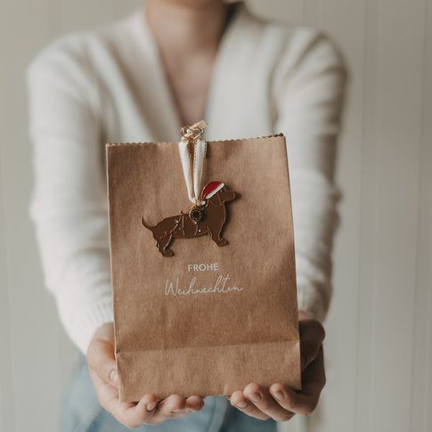 Person hält weihnachtliche Kraftpapier-Geschenktüte mit „Frohe Weihnachten“ und Dackel-Anhänger