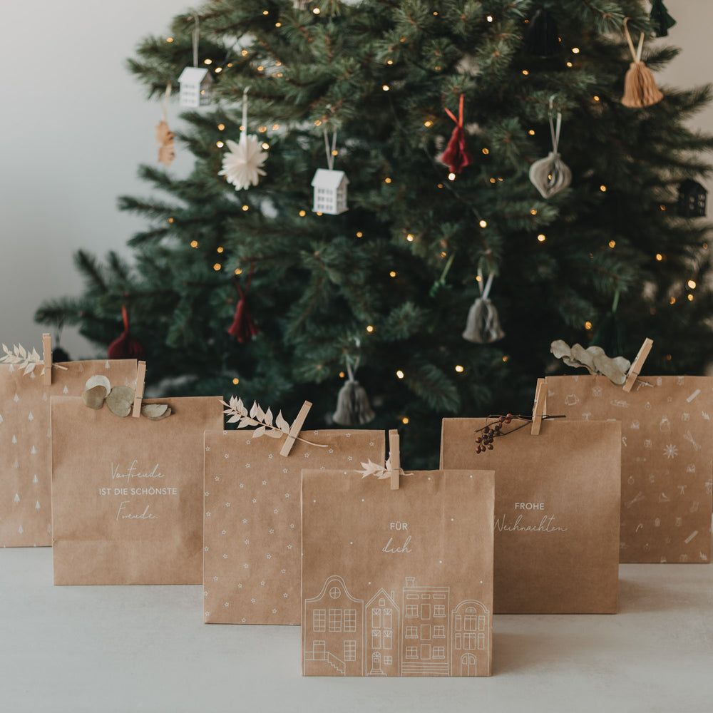 Weihnachtliche Kraftpapier-Geschenktüten mit verschiedenen weißen Motiven vor beleuchtetem Tannenbaum