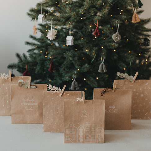 Weihnachtliche Kraftpapier-Geschenktüten mit verschiedenen weißen Motiven vor beleuchtetem Tannenbaum