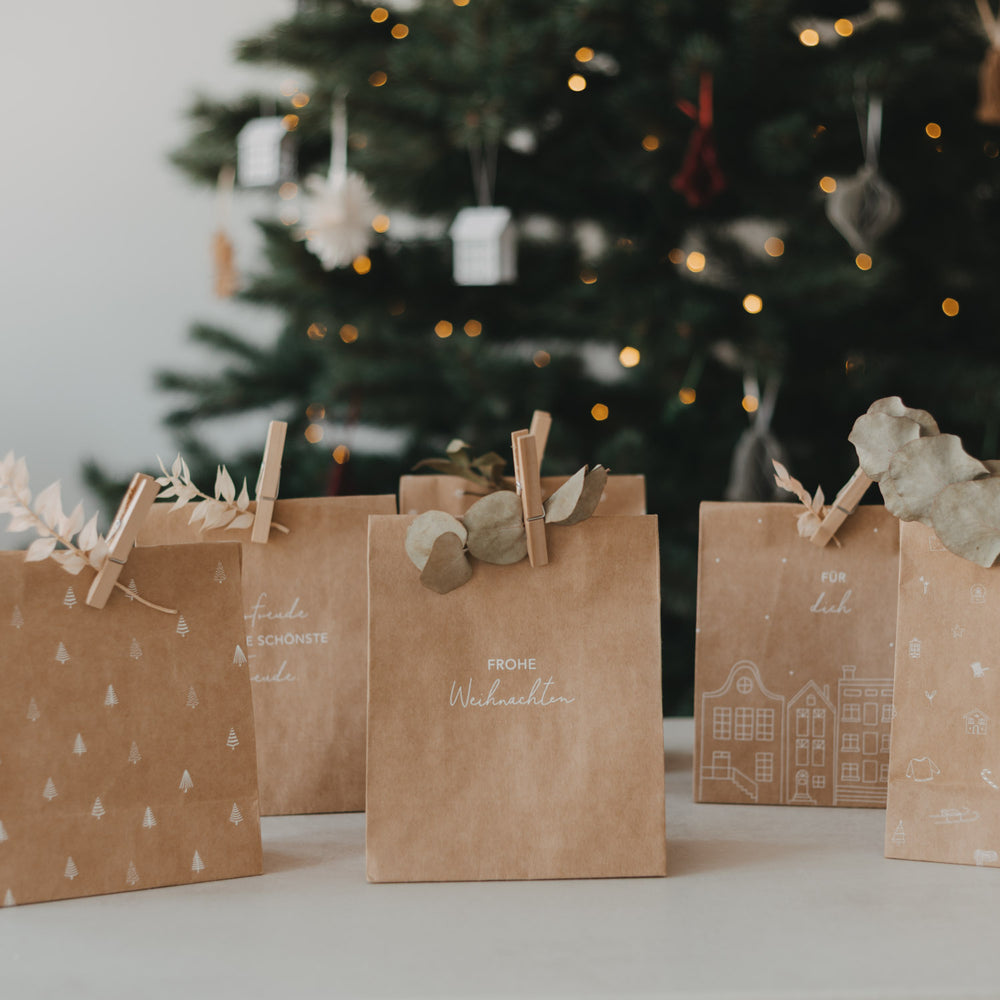 Weihnachtliche Geschenktüten aus Kraftpapier mit weißen Motiven vor geschmücktem Weihnachtsbaum