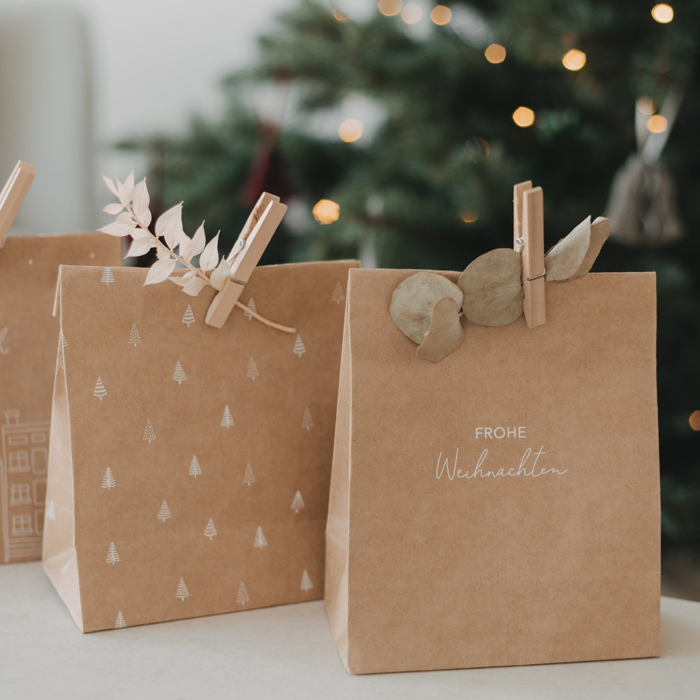 Geschenktüten aus Kraftpapier mit „Frohe Weihnachten“ und Tannenbaum-Motiv vor Weihnachtsbaum