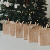 Dekorierte Geschenktüten aus Kraftpapier mit Weihnachtsmotiven und Schriftzügen vor Weihnachtsbaum