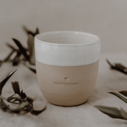 Ein einzelner Becher mit dem Schriftzug CIAO KAKAO steht auf einem beigen Leinentuch, umrahmt von Olivenzweigen.