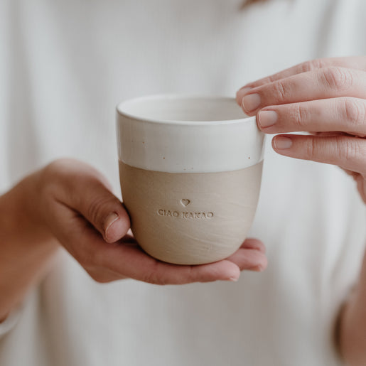 Zwei Hände halten einen Becher mit matter, zweifarbiger Keramik in Beige und Weiß sowie dem Schriftzug CIAO KAKAO vor einem hellen, neutralen Hintergrund.